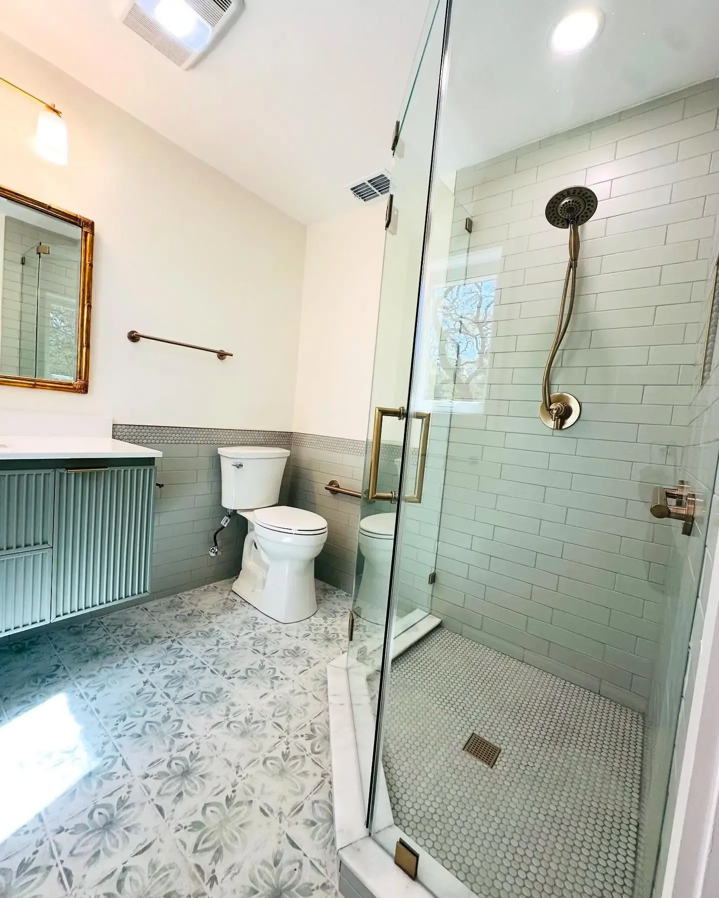 Modern bathroom with a glass shower enclosure, a toilet, a mint green vanity with a sink, a mirror with a gold frame, and patterned tile flooring.