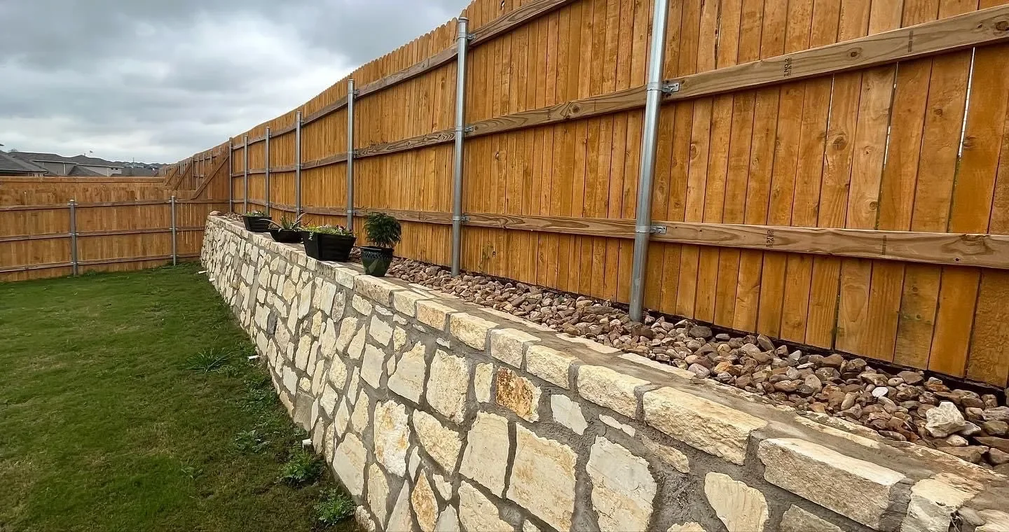 A backyard with a stone retaining wall topped with a row of potted plants, a wooden privacy fence, and a green lawn on a cloudy day.