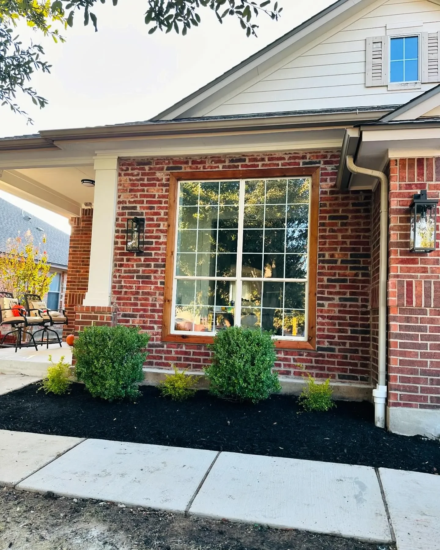 Front view of a house with a brick exterior, a large window with wooden trim, two black lantern-style wall lights, and landscaped bushes in front, with a concrete sidewalk in the foreground.