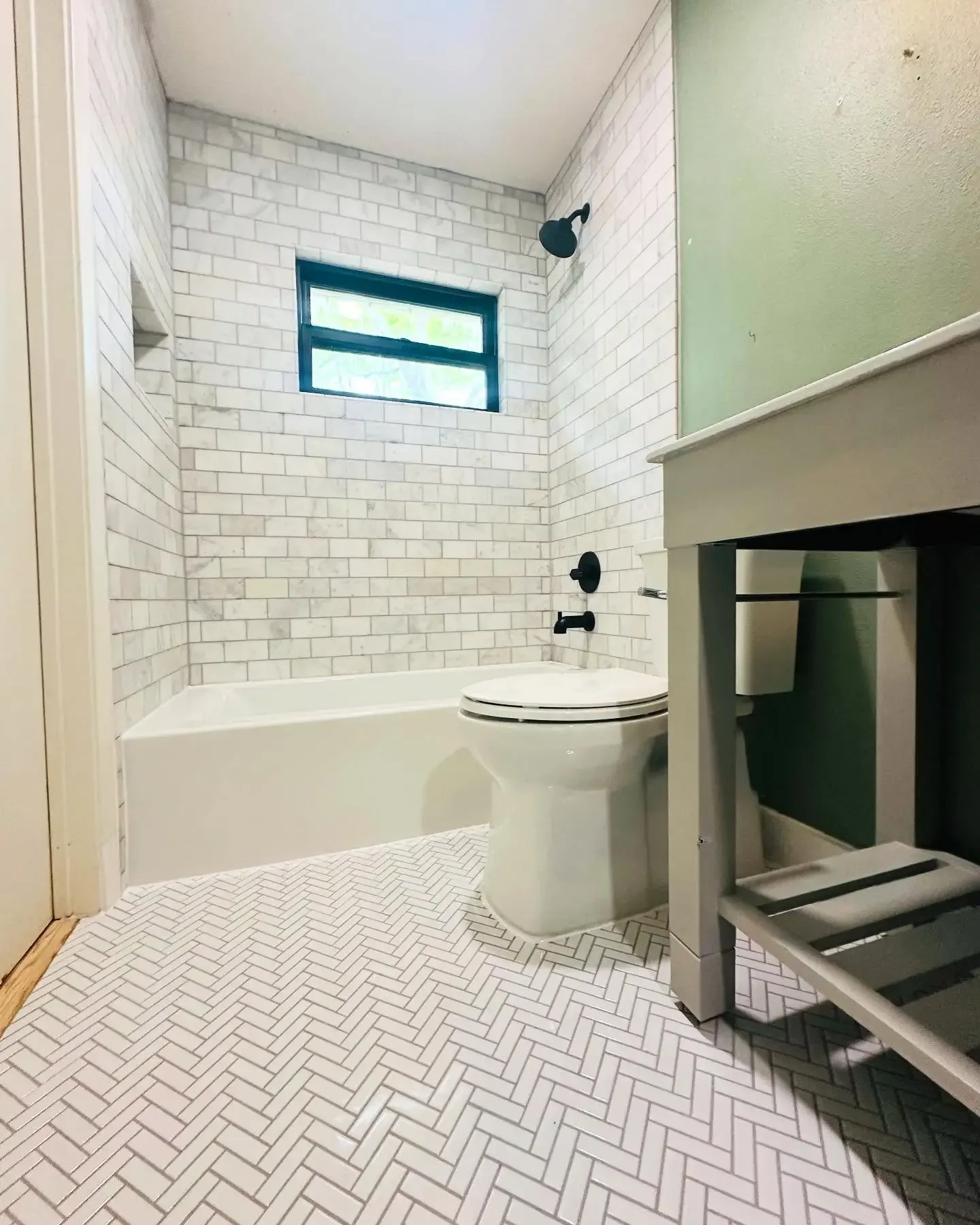 Modern bathroom with white herringbone tile floor, white brick wall behind bathtub, black fixtures, small window, and green wall.