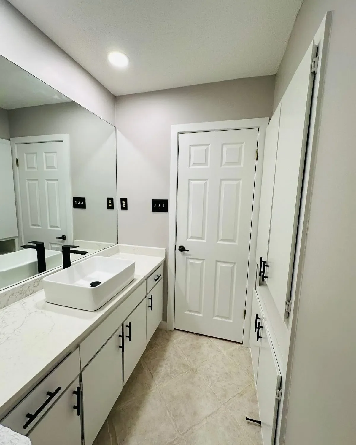 Modern bathroom with white cabinetry, a vessel sink, black fixtures, large mirror, and neutral tile flooring.