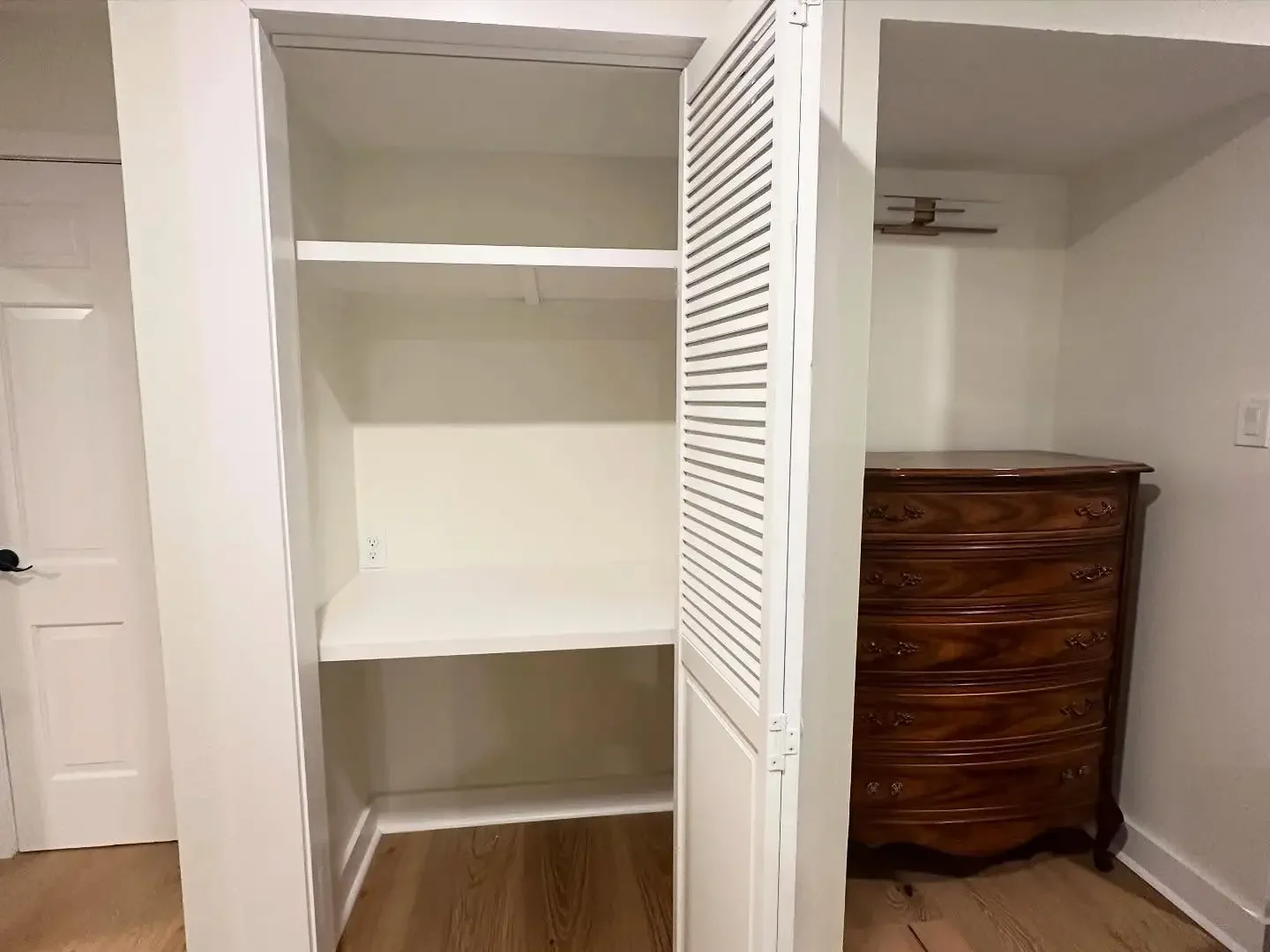 Empty built-in closet with white shelves and doors, next to a wooden dresser in a room with hardwood floor and white walls.