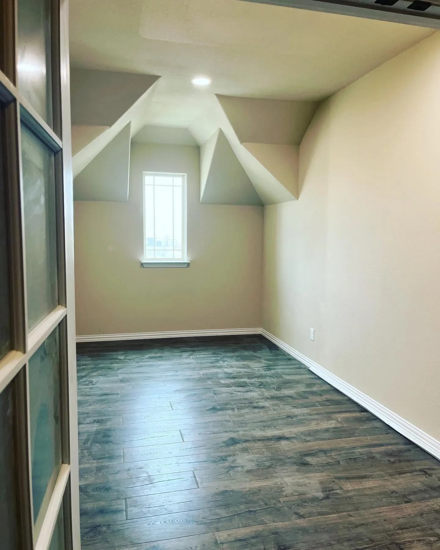 Empty room with beige walls, dark wood flooring, a small window, and angled ceiling panels.