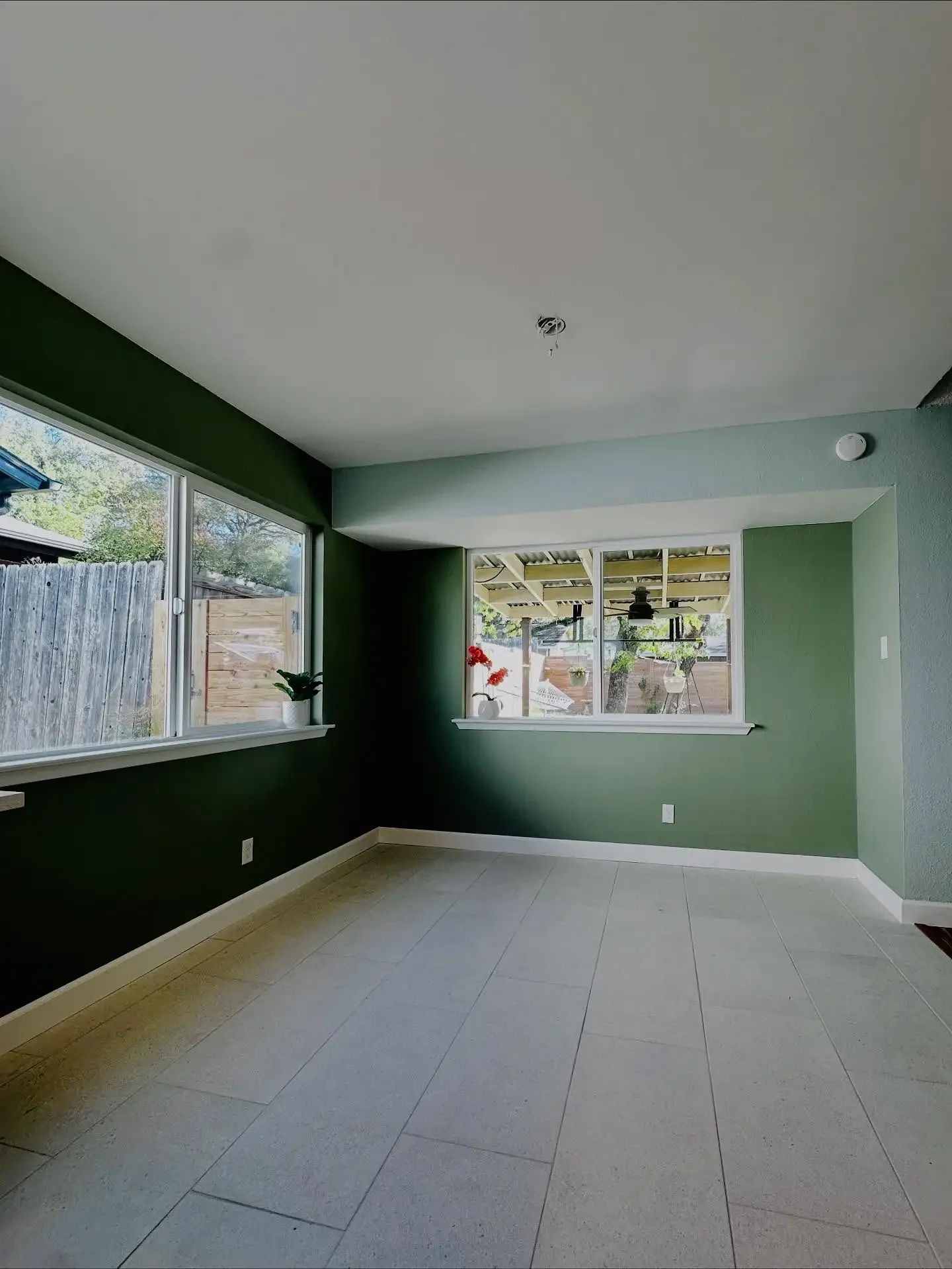 Empty room with large windows, light-colored tile floor, green walls, and a ceiling fixture with wires hanging. Outside view shows a covered patio with hanging plants and a tree.