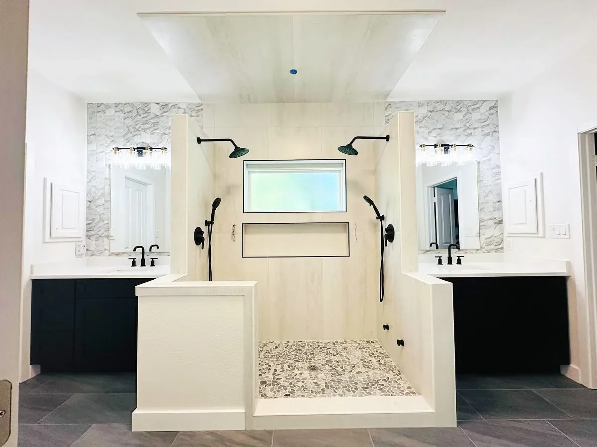 Modern dual vanity bathroom with a shower in the center, featuring two black showerheads and handheld shower fixtures, large mirrors with light fixtures, white walls, and dark cabinetry.