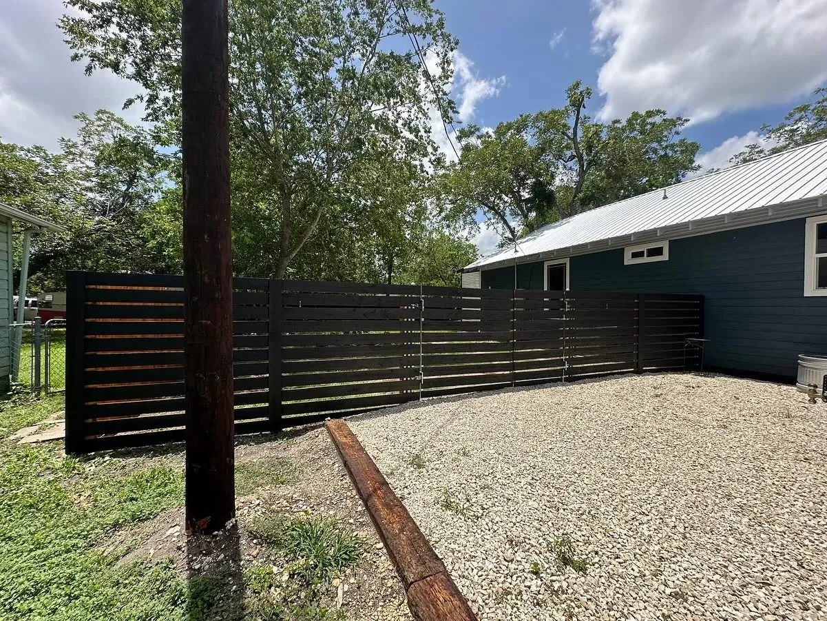 A backyard with a modern black horizontal wooden fence, trees in the background, a gravel ground, and part of a blue house with a metal roof.