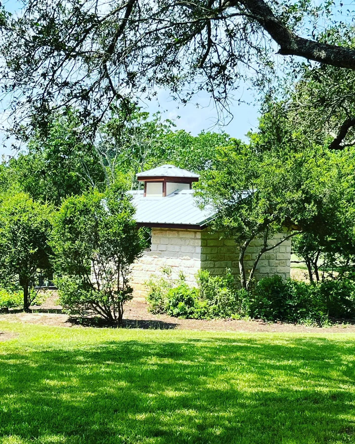 A small stone building with a metal roof, surrounded by lush green trees and bushes, under a partly cloudy sky.