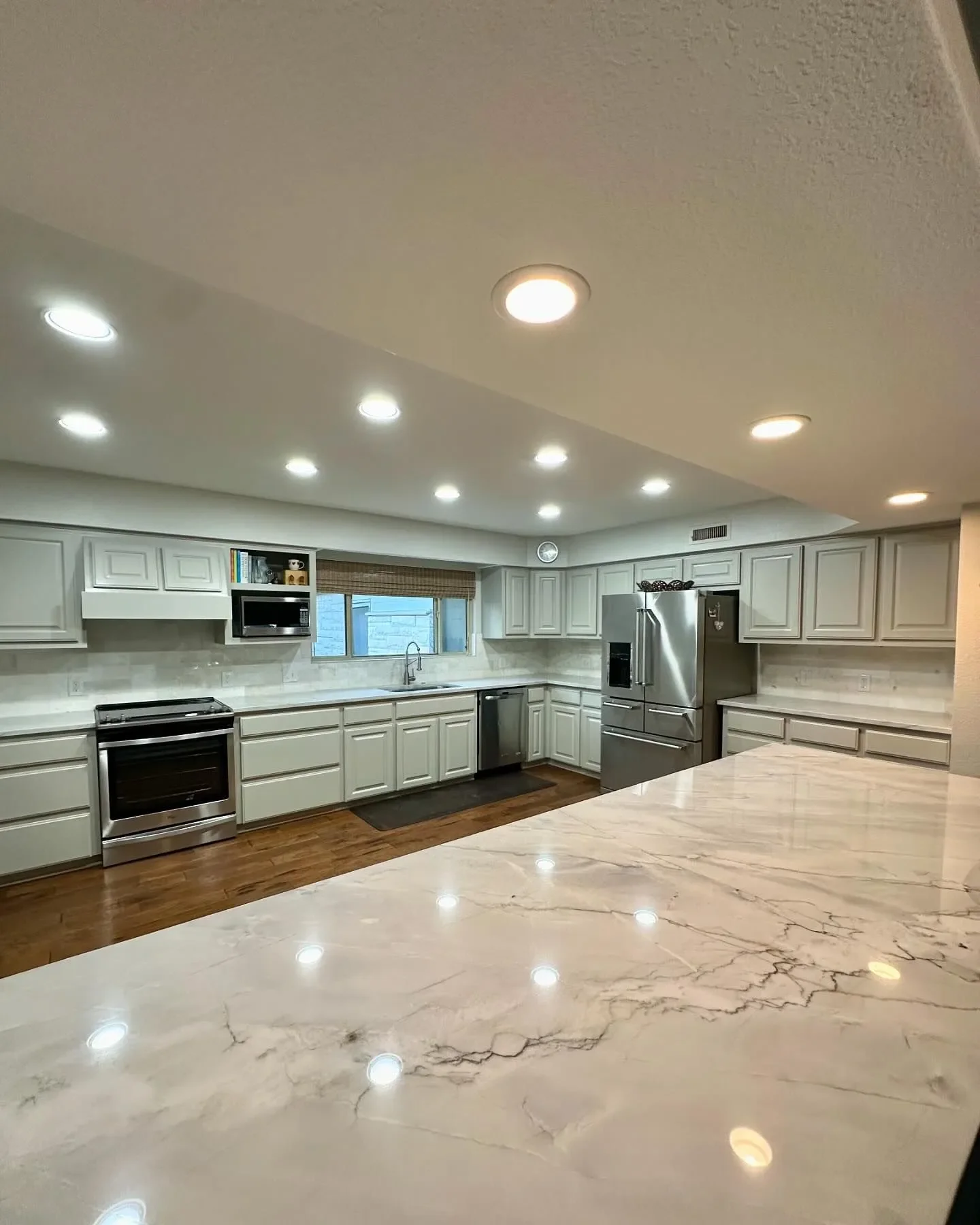 Modern kitchen with white cabinets, stainless steel appliances, marble countertop, and wooden flooring, illuminated by recessed ceiling lights.