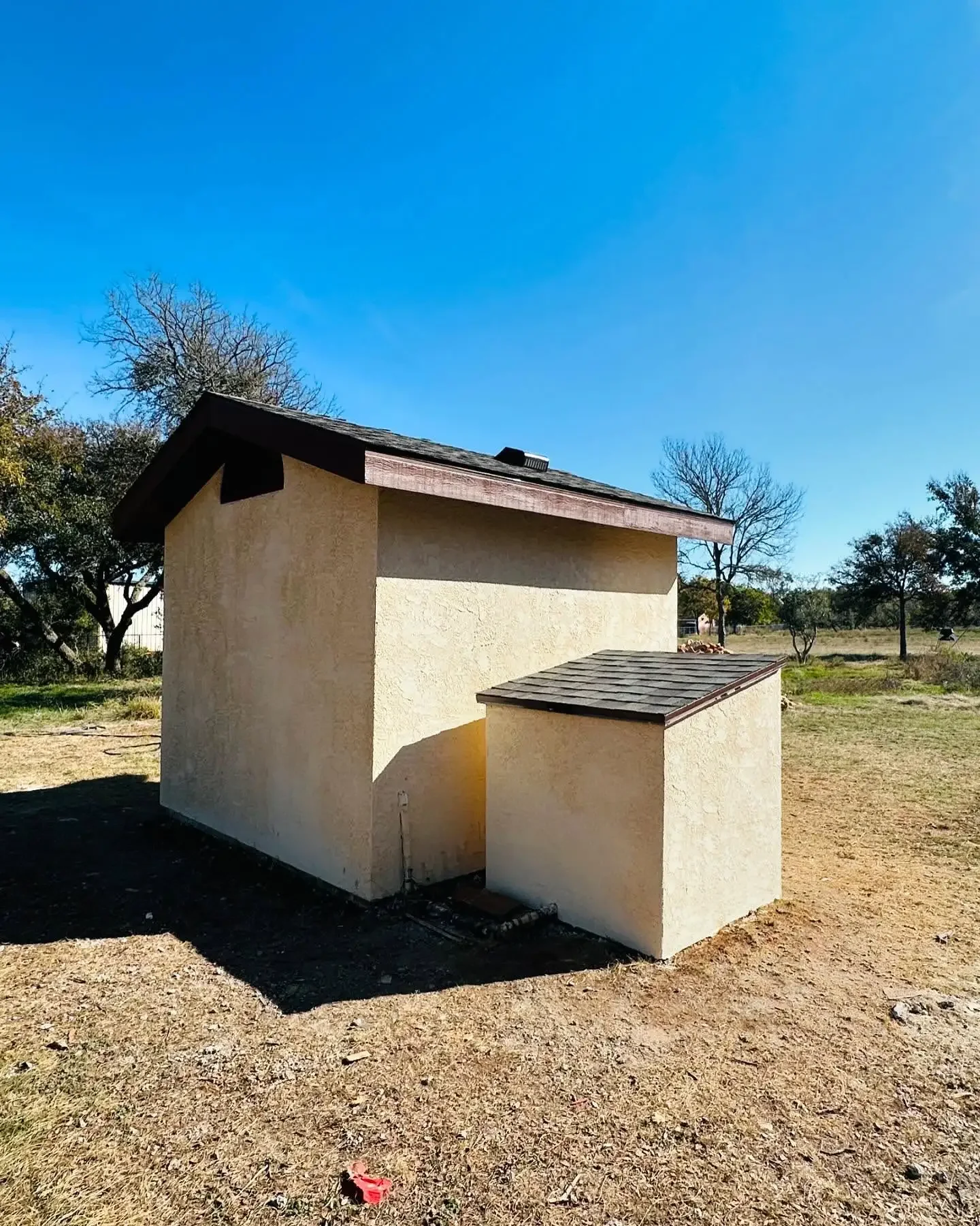 Small beige building with a sloped roof, situated outdoors with trees and a clear blue sky in the background.