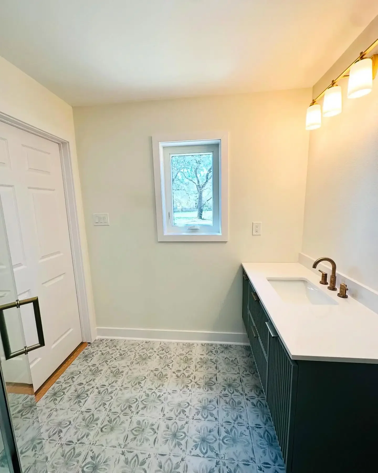 A bathroom with a small window showing trees outside, a dark vanity with a white countertop and a bronze faucet, a three-light fixture above, and patterned floor tiles.