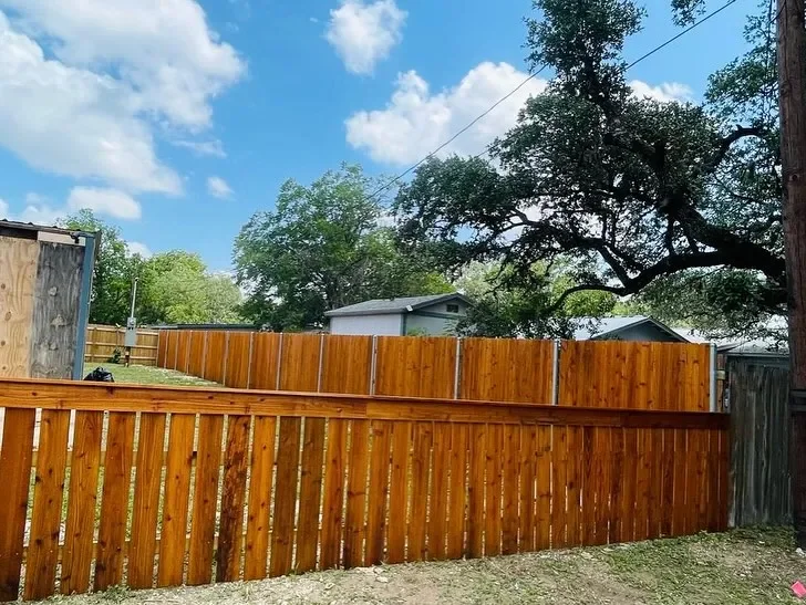 Side view of a backyard with a newly installed wooden fence, green grass, a tree, and a small gray building in the background under a partly cloudy sky.