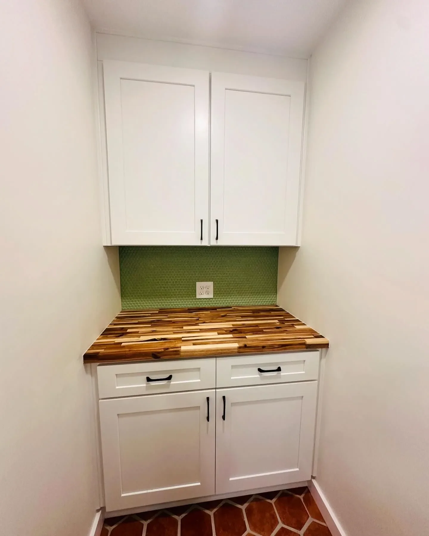A small corner cabinet setup with white upper and lower cabinets, a wooden countertop, and a green textured backsplash, in a room with cream walls and reddish-brown tiled flooring.