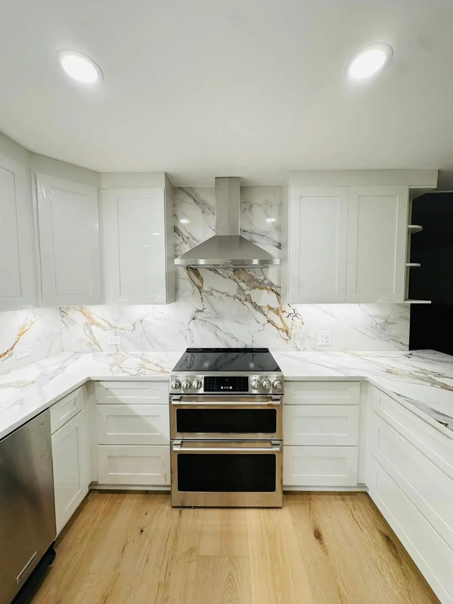 Modern kitchen with white cabinetry, marble backsplash and countertops, stainless steel oven and range hood, hardwood floor.