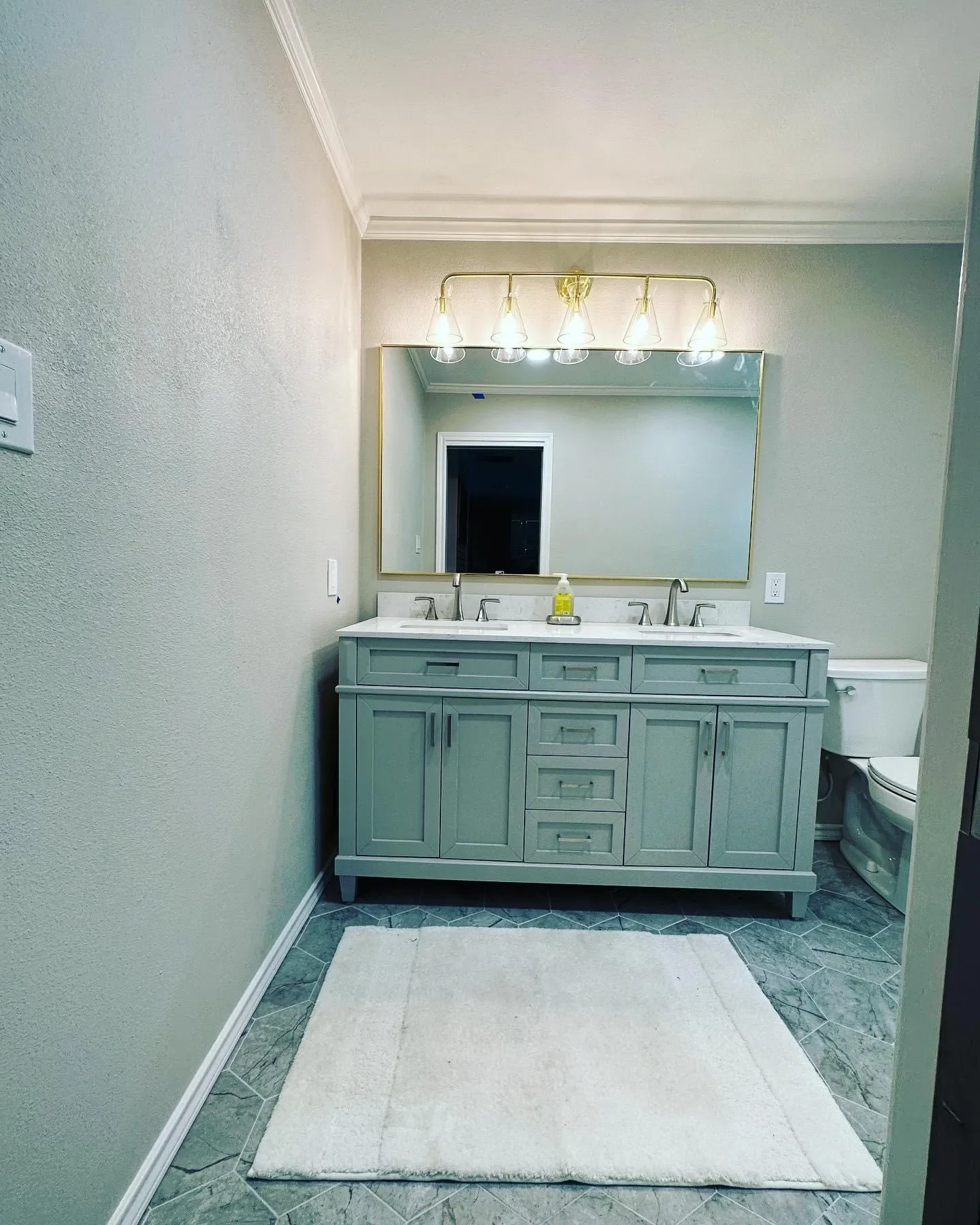 Bathroom with light green vanity, large mirror, four light fixtures, white countertop, two sinks, soap dispenser, toilet in the background, and a beige rug on dark gray tiled floor.