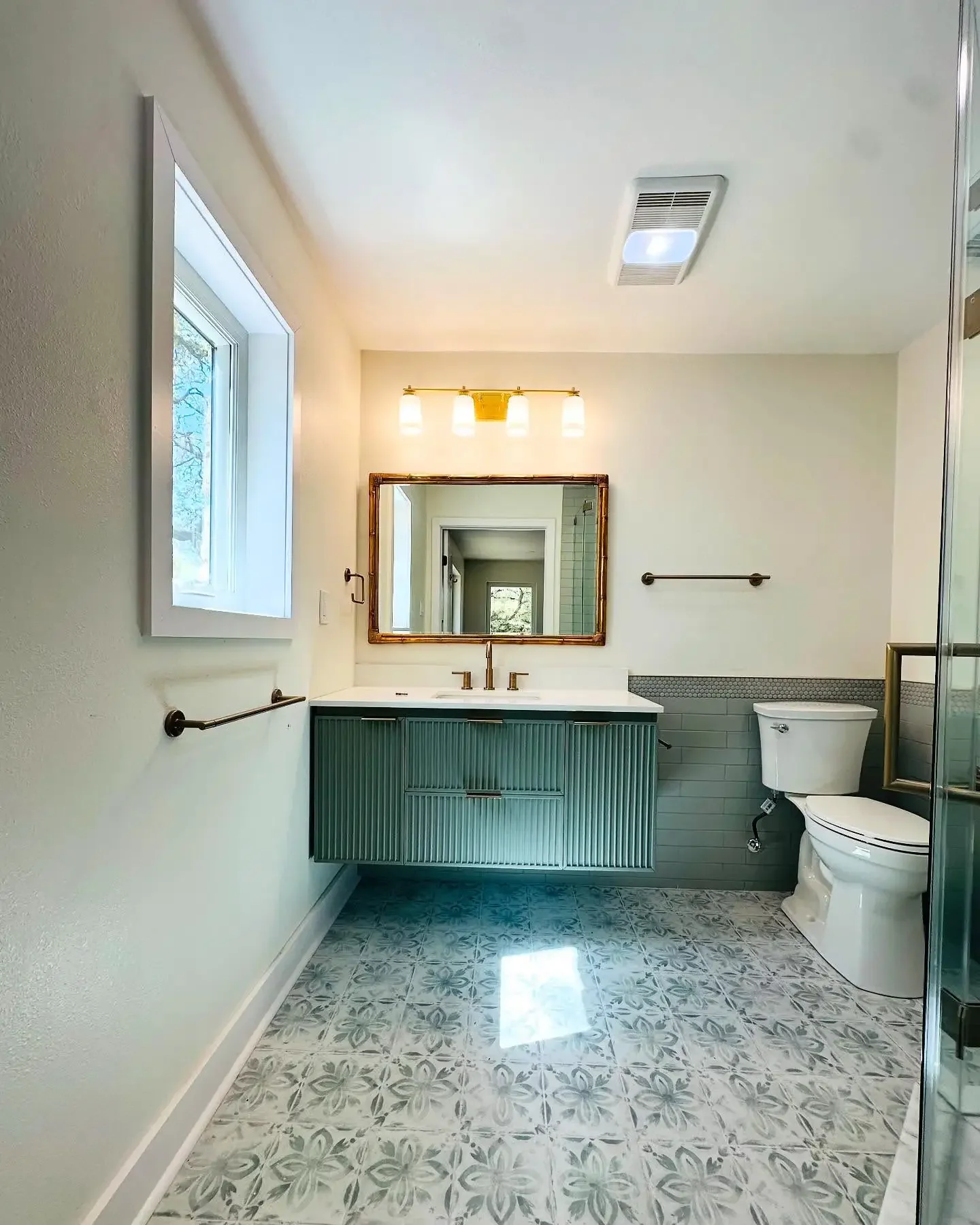 A modern bathroom with a wall-mounted vanity with a green textured finish, a white countertop, and a rectangular mirror above. There is a window on the left, a light fixture with four bulbs above the mirror, a toilet on the right, and a shower enclos