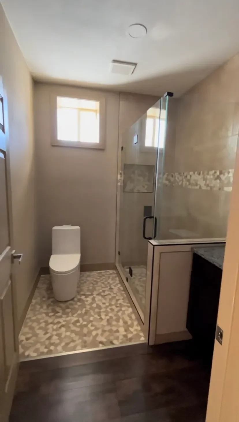 Small bathroom with a modern toilet, a glass-enclosed shower, and a window bringing in natural light. Light-colored tiles and dark wooden flooring.