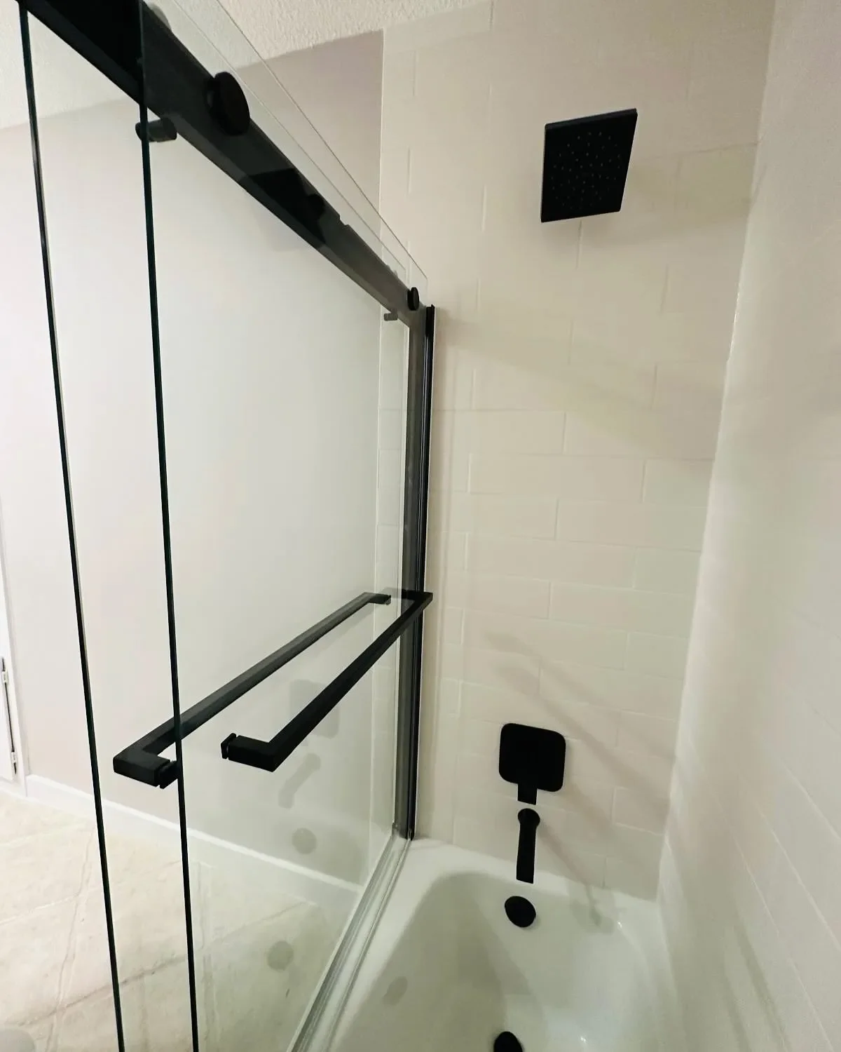 A modern bathroom shower with a glass enclosure, black fixtures including a showerhead, a handle, and black support bars, with a white tiled wall and a bathtub.