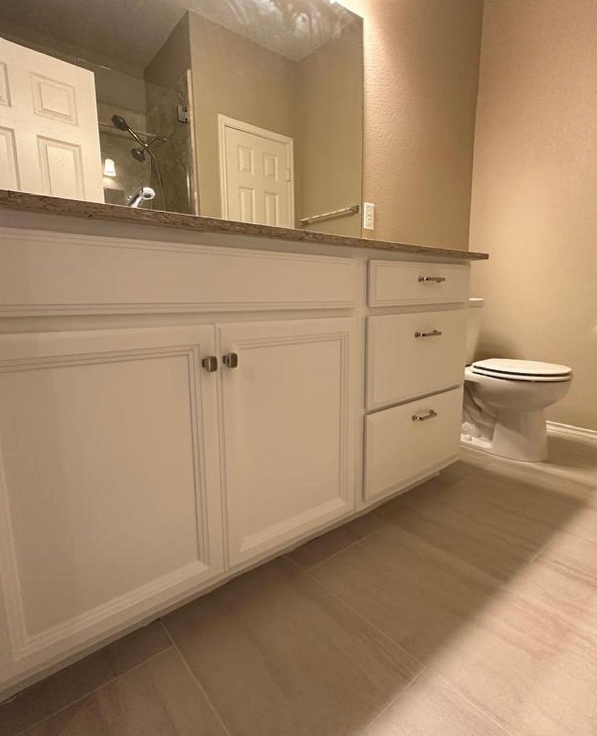 Bathroom with white cabinetry, granite countertop, and a toilet. There is a mirror above the sink and a shower visible in the reflection.