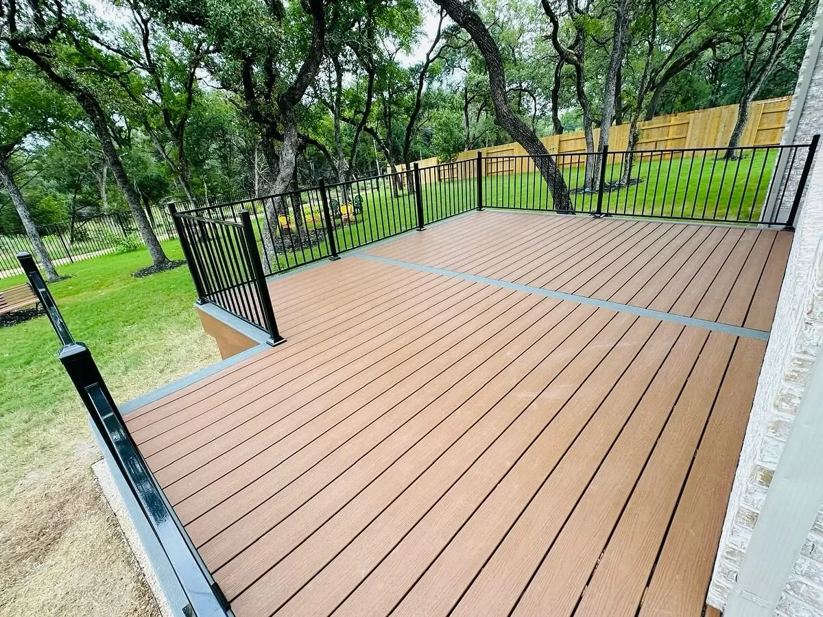 A wooden deck with black metal railing in a backyard with green grass, trees, and a wooden fence.