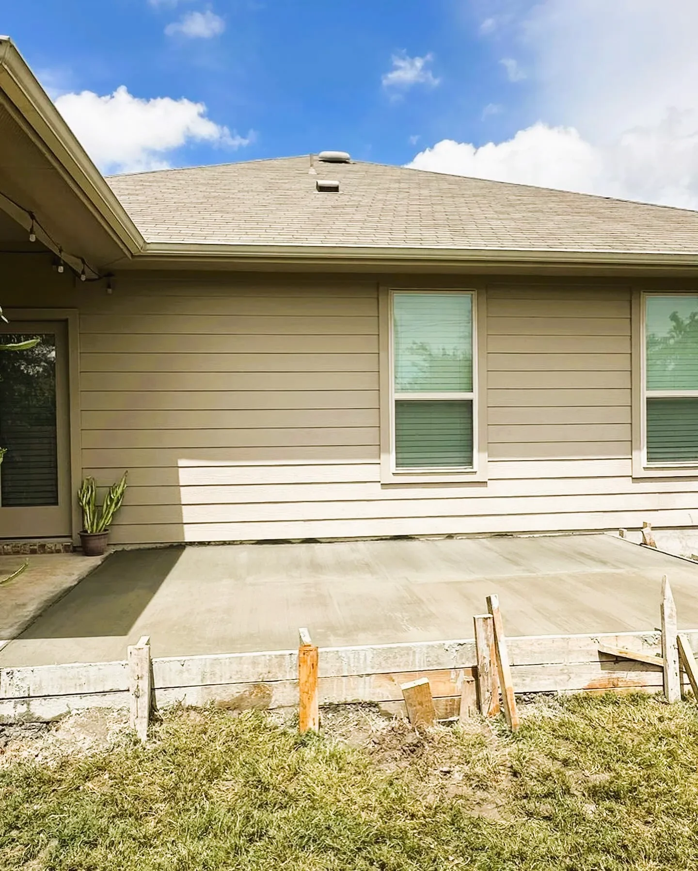 Newly poured concrete patio in backyard with wooden formwork and some grass in front, beige house with two windows and a plant in a pot, blue sky with clouds.