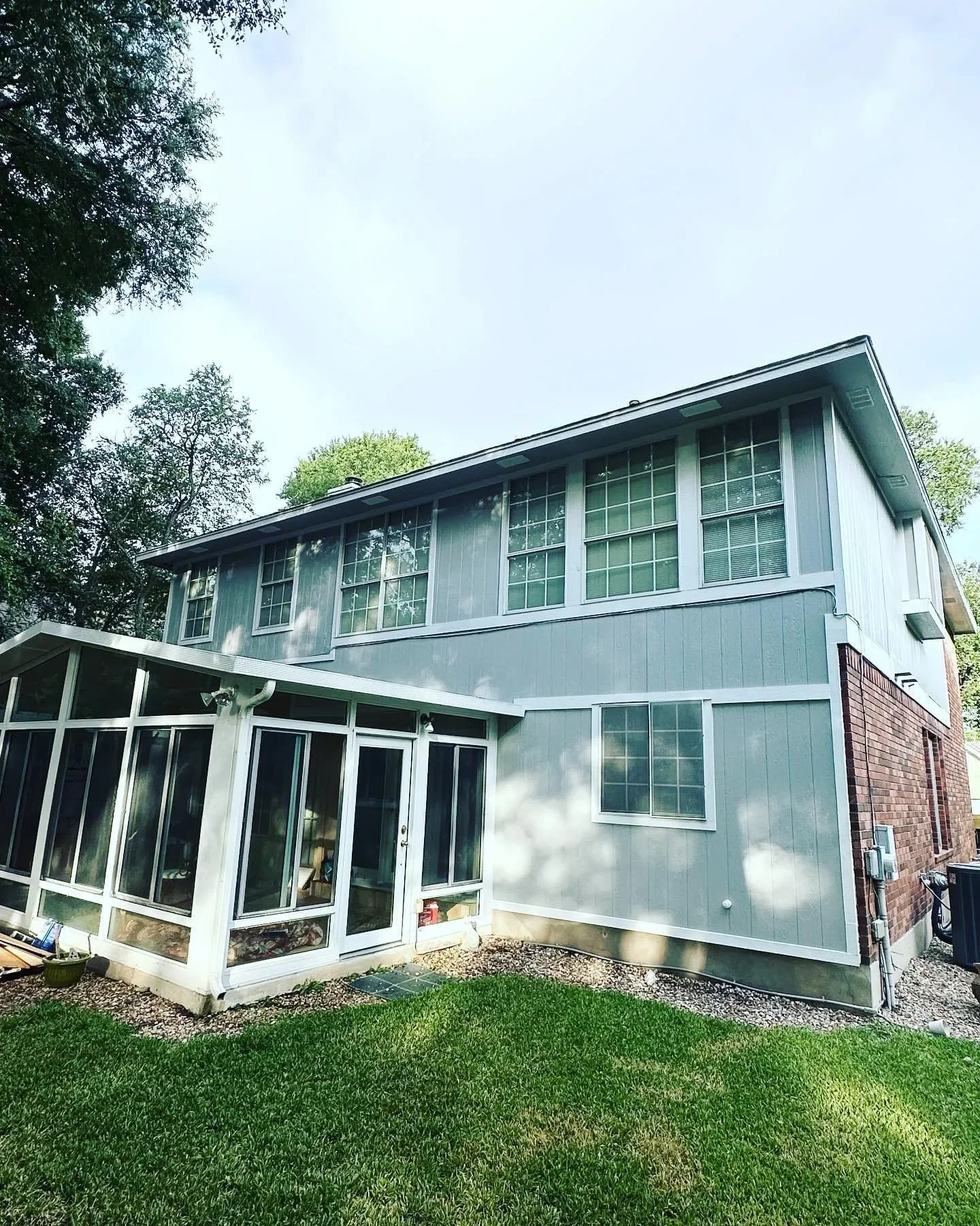 Backyard of a two-story house with green siding, multiple windows, and a sunroom with glass walls, surrounded by a green lawn.
