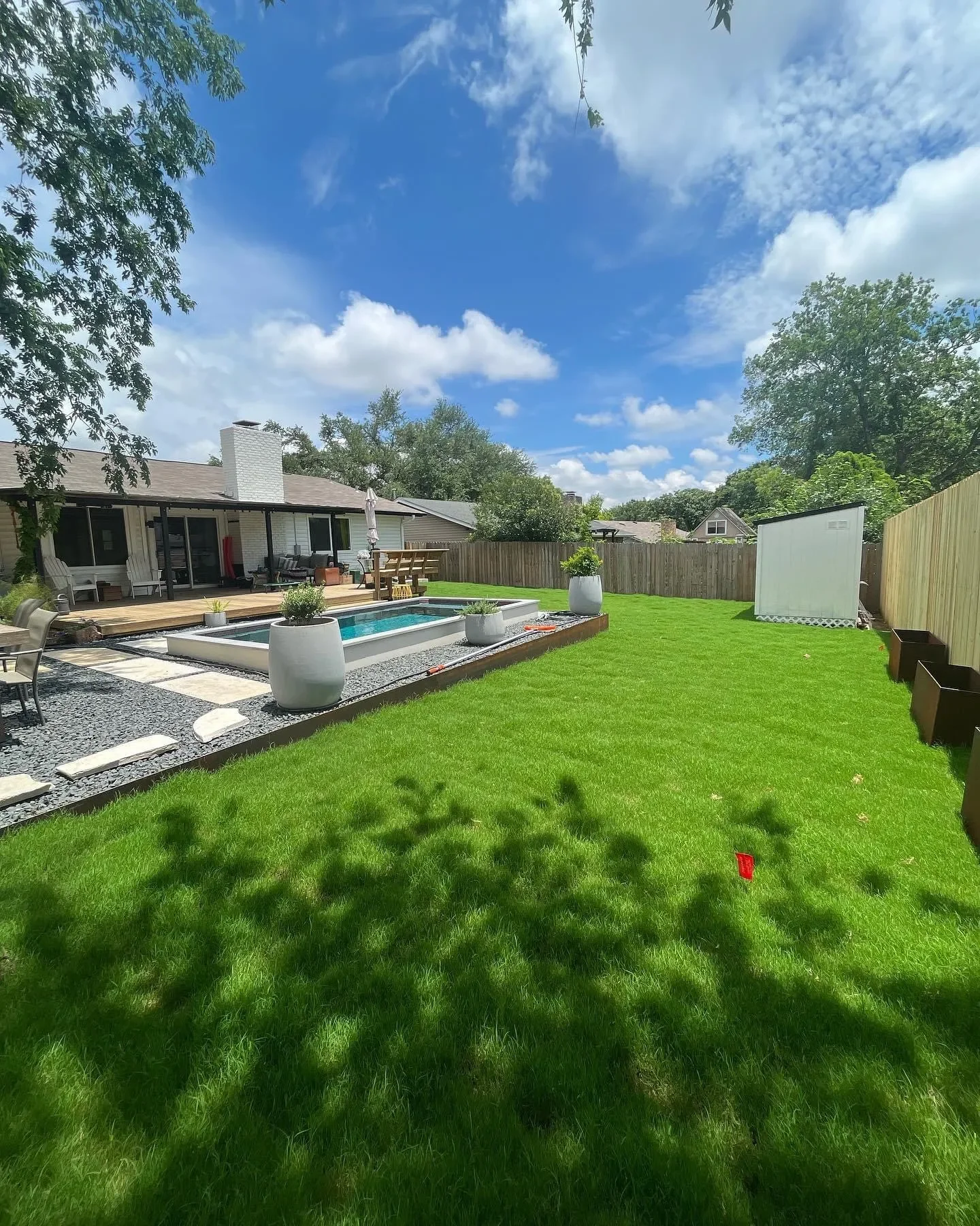 A backyard with a well-maintained green lawn, a small pool with surrounding potted plants, a sitting area, and a wooden fence under a partly cloudy sky.