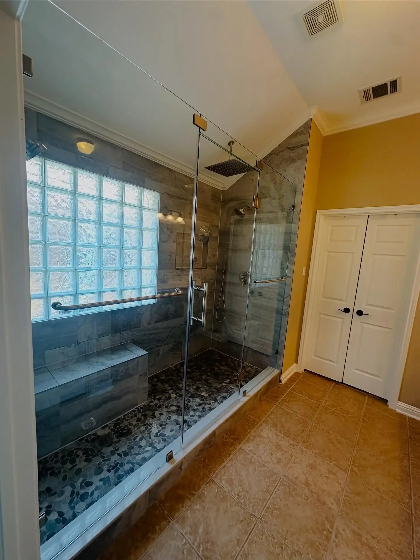 Bathroom with a glass-enclosed shower, pebble stone floor, gray and beige tile walls, a window with glass blocks, a bench inside, and a double-door closet on the right wall.
