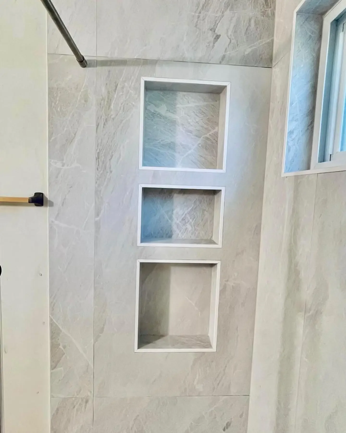 A modern shower with beige marble tiles, three built-in shelves, a grab bar, and a small window.