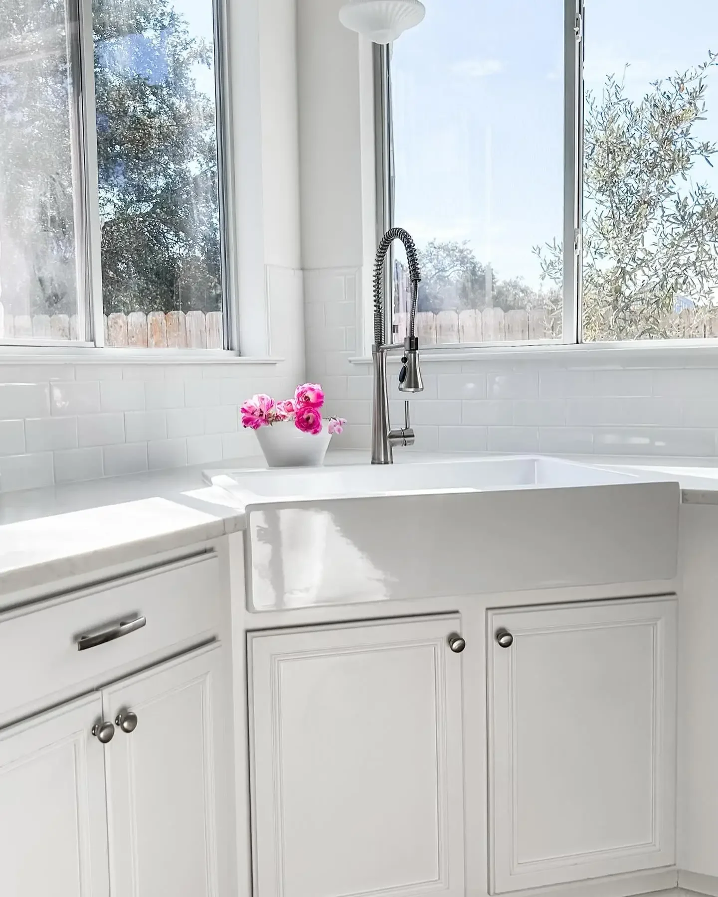 Bright kitchen with a white farmhouse sink, chrome faucet, and white cabinetry, featuring a window with a view of trees and a pot of pink flowers on the countertop.