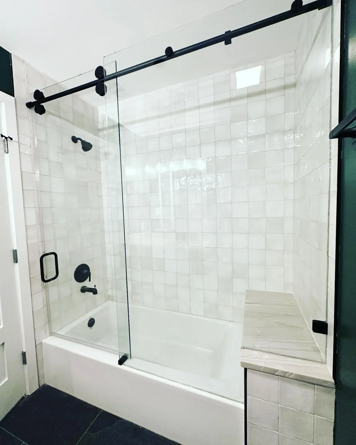A modern walk-in shower with a glass door, black fixtures, white tiled walls, a built-in marble bench, and dark tiled flooring.