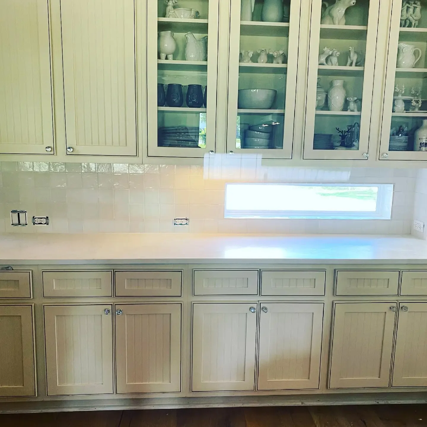 Kitchen with beige cabinets, glass-front upper cabinets filled with white and dark-colored dishes, and a horizontal window above the countertop.