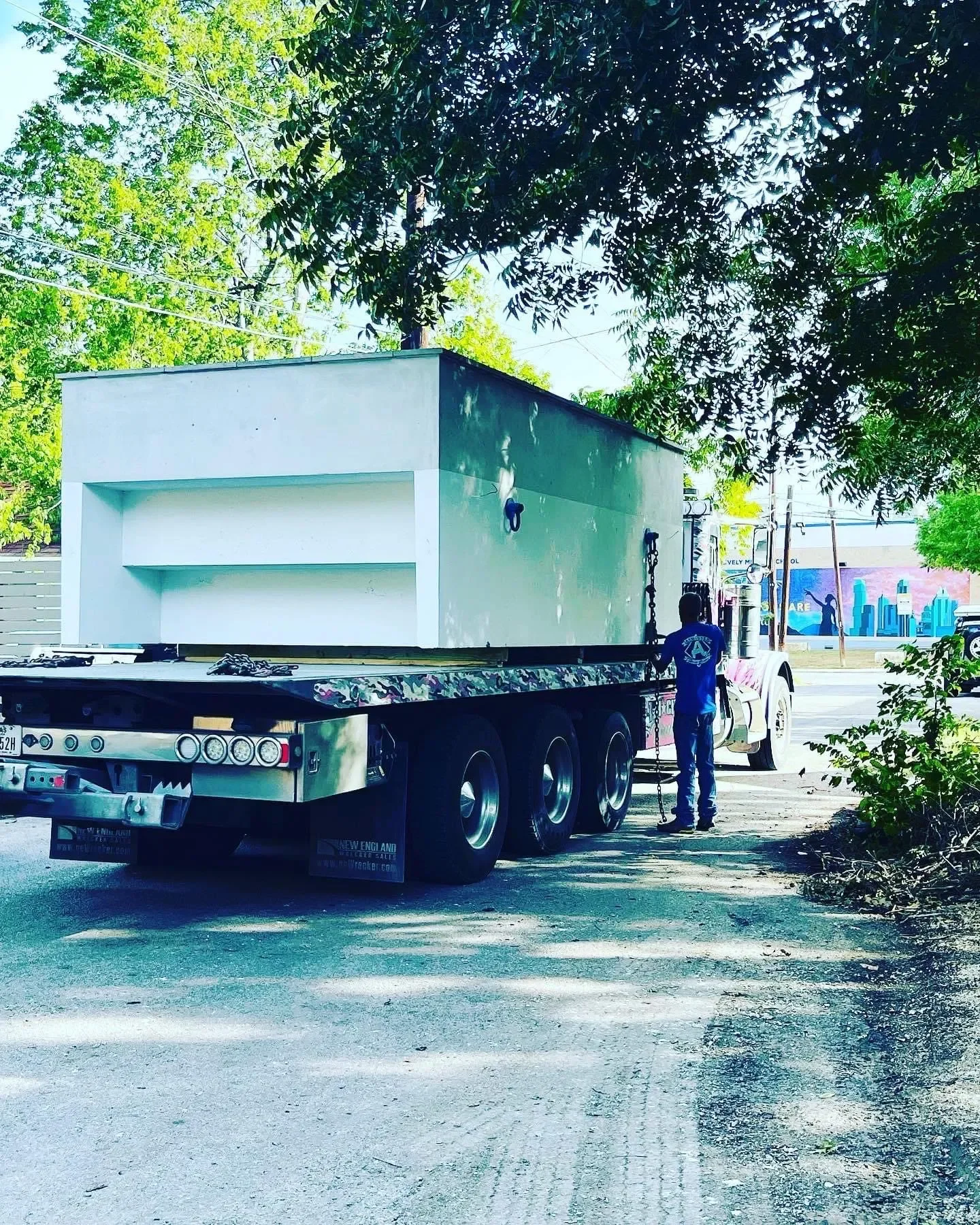 A person standing on the street near a flatbed truck carrying a large, white rectangular structure with two shelves, surrounded by trees and urban environment.