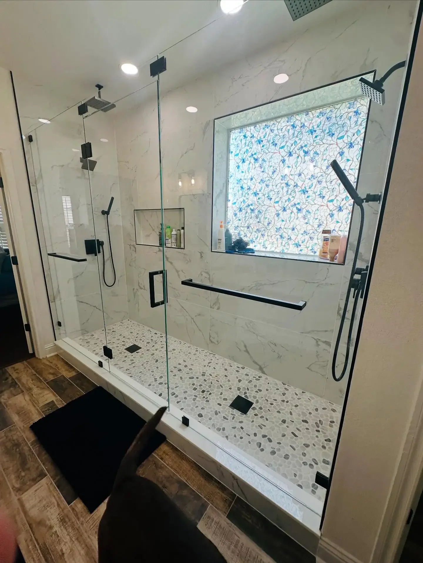 A modern walk-in shower in a bathroom with large marble tiles and a pebble floor. It has a glass enclosure, two black showerheads, a built-in niche for toiletries, and a window with a colorful floral-patterned blind.