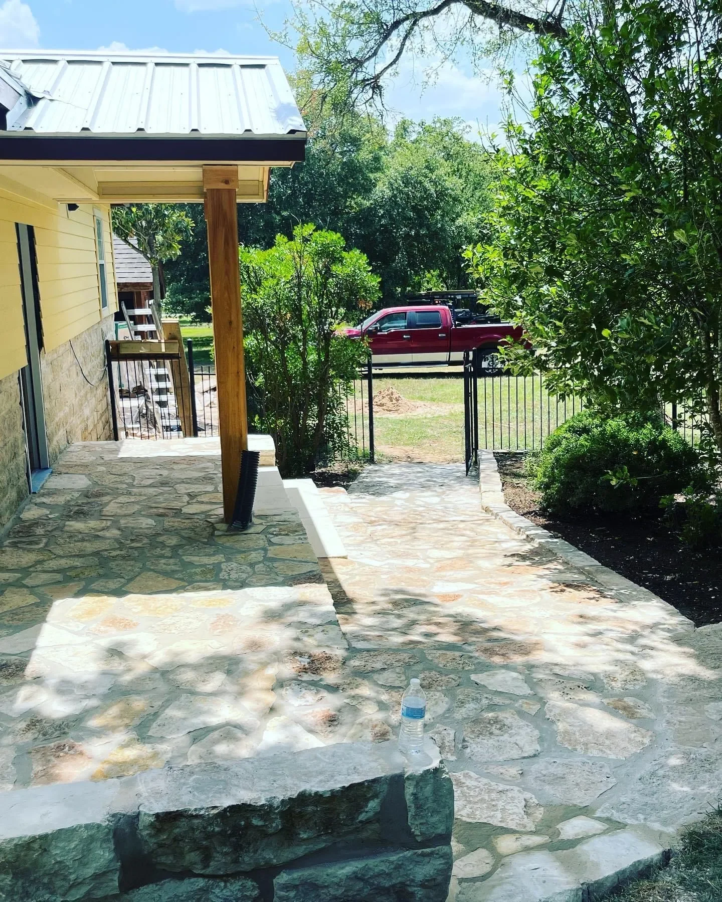 Stone patio and walkway outside a yellow house with a metal roof, greenery, a red pickup truck parked on the grass, and a black metal gate.