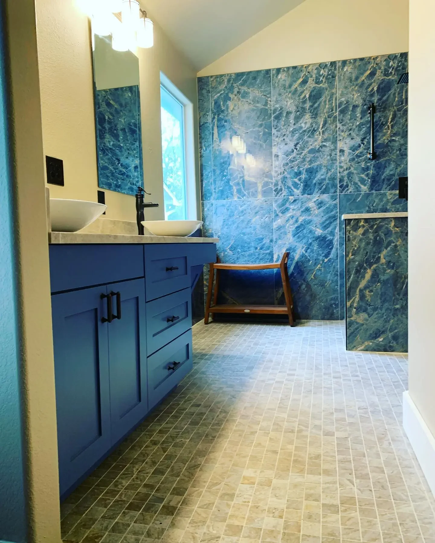 A modern bathroom with a blue vanity with drawers, a white countertop, and two vessel sinks with black faucets. There is a mirror above the vanity and a window bringing in natural light. The wall behind the vanity is painted a light color, and there 