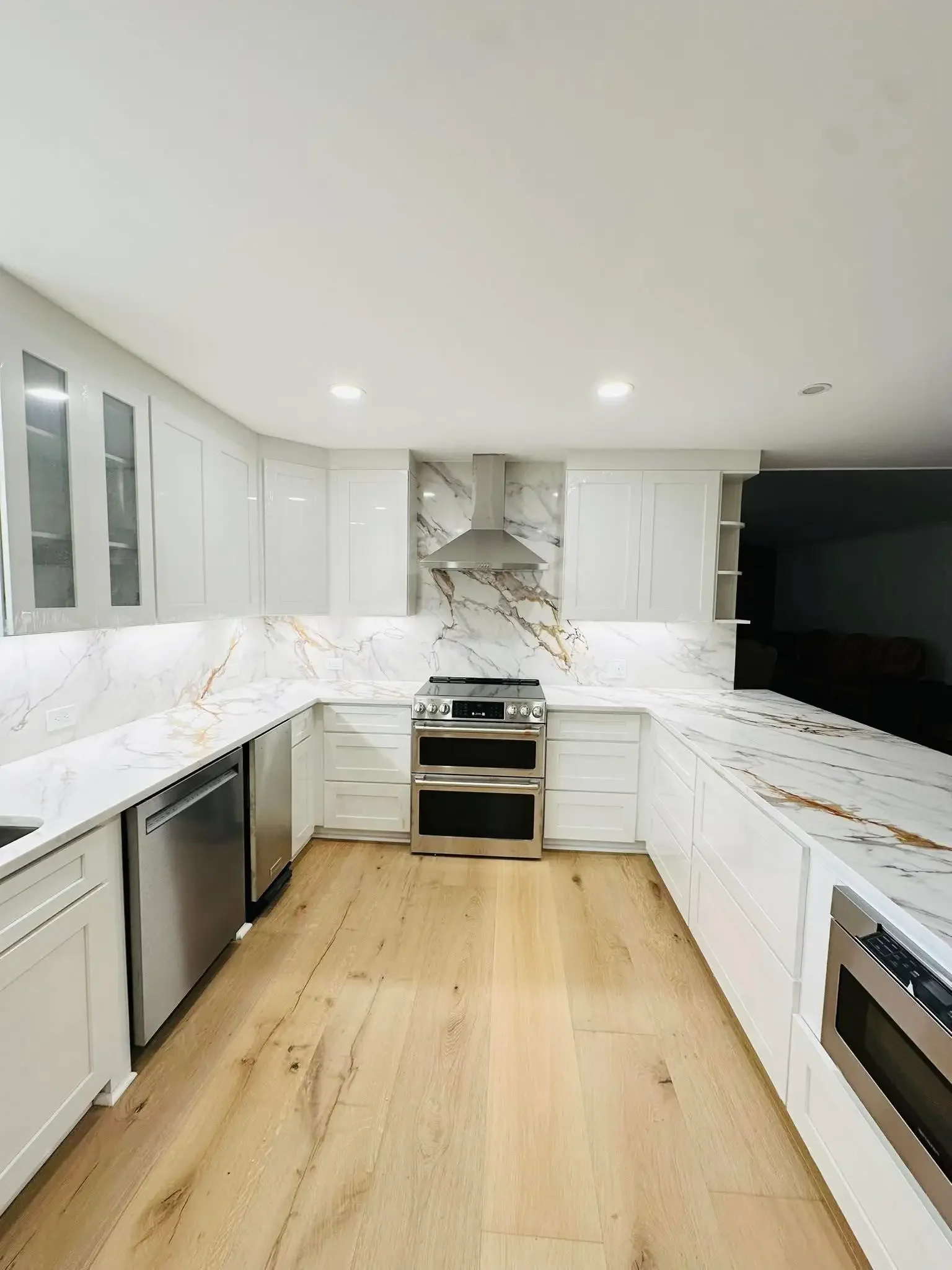 Modern kitchen with white cabinets, marble countertops, and stainless steel appliances, including oven and microwave, with a light wooden floor.