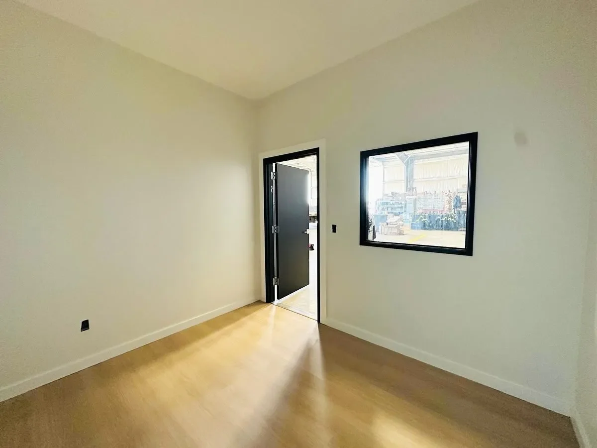 Empty room with light-colored walls, wooden floor, black door slightly open, and a window showing an industrial outdoor scene.