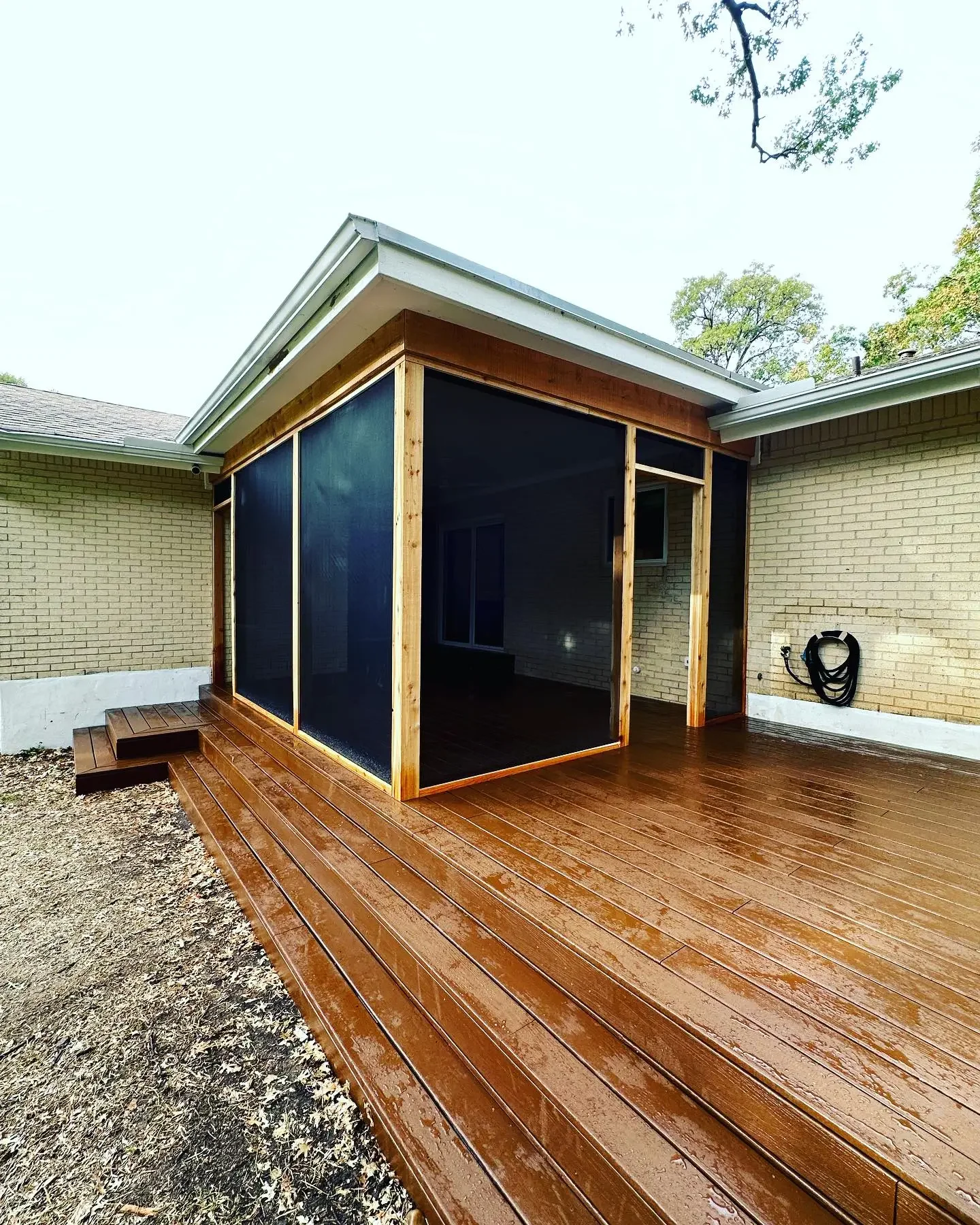 Backyard with a newly built, screened-in porch made of wood and glass, adjacent to a brick house. The wooden deck is wet, indicating recent rain, and outdoor trees are visible overhead.