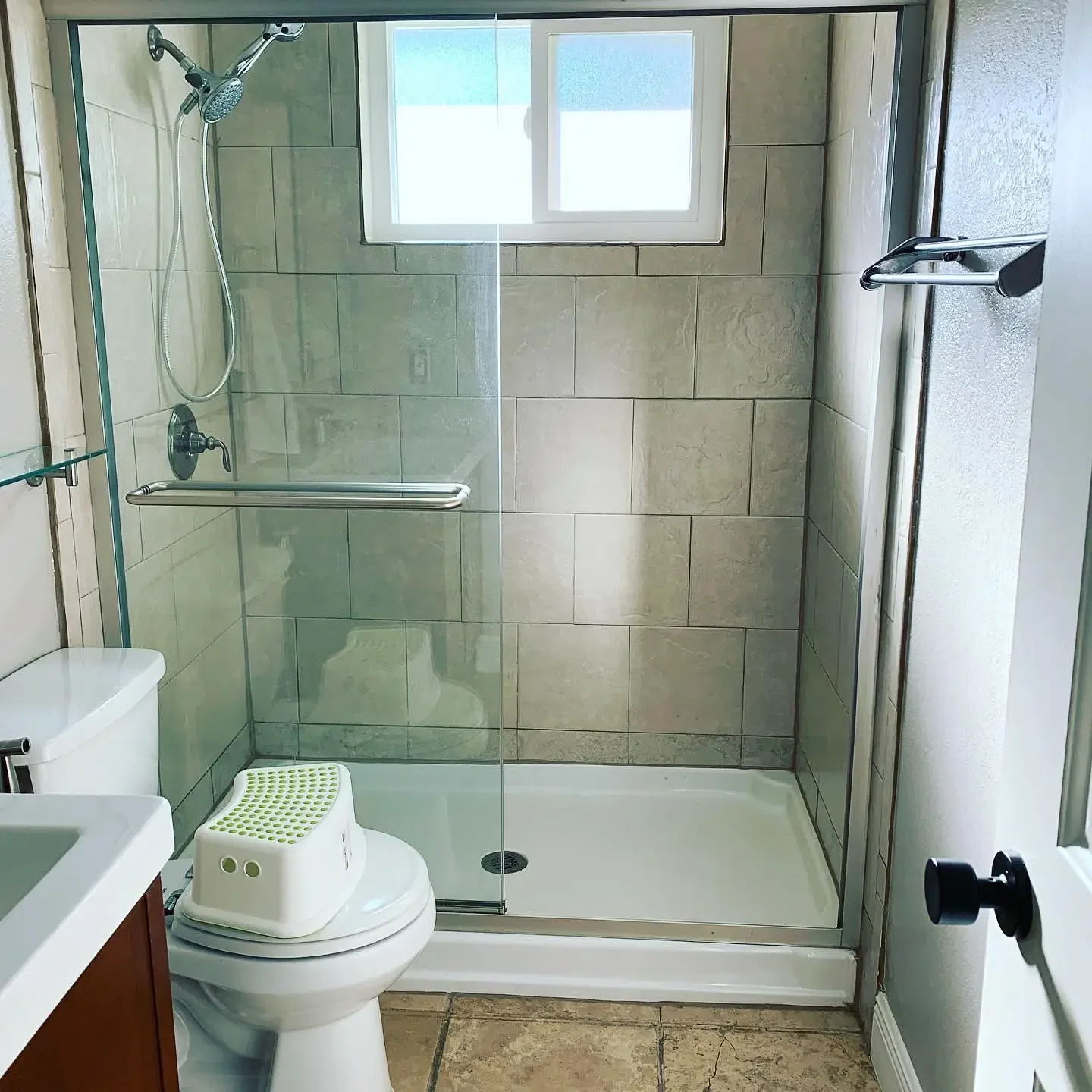 A bathroom with a walk-in shower enclosed by glass, a window in the shower, a white toilet with a baby bath seat on top, part of a sink with a wooden cabinet, and a tiled floor.