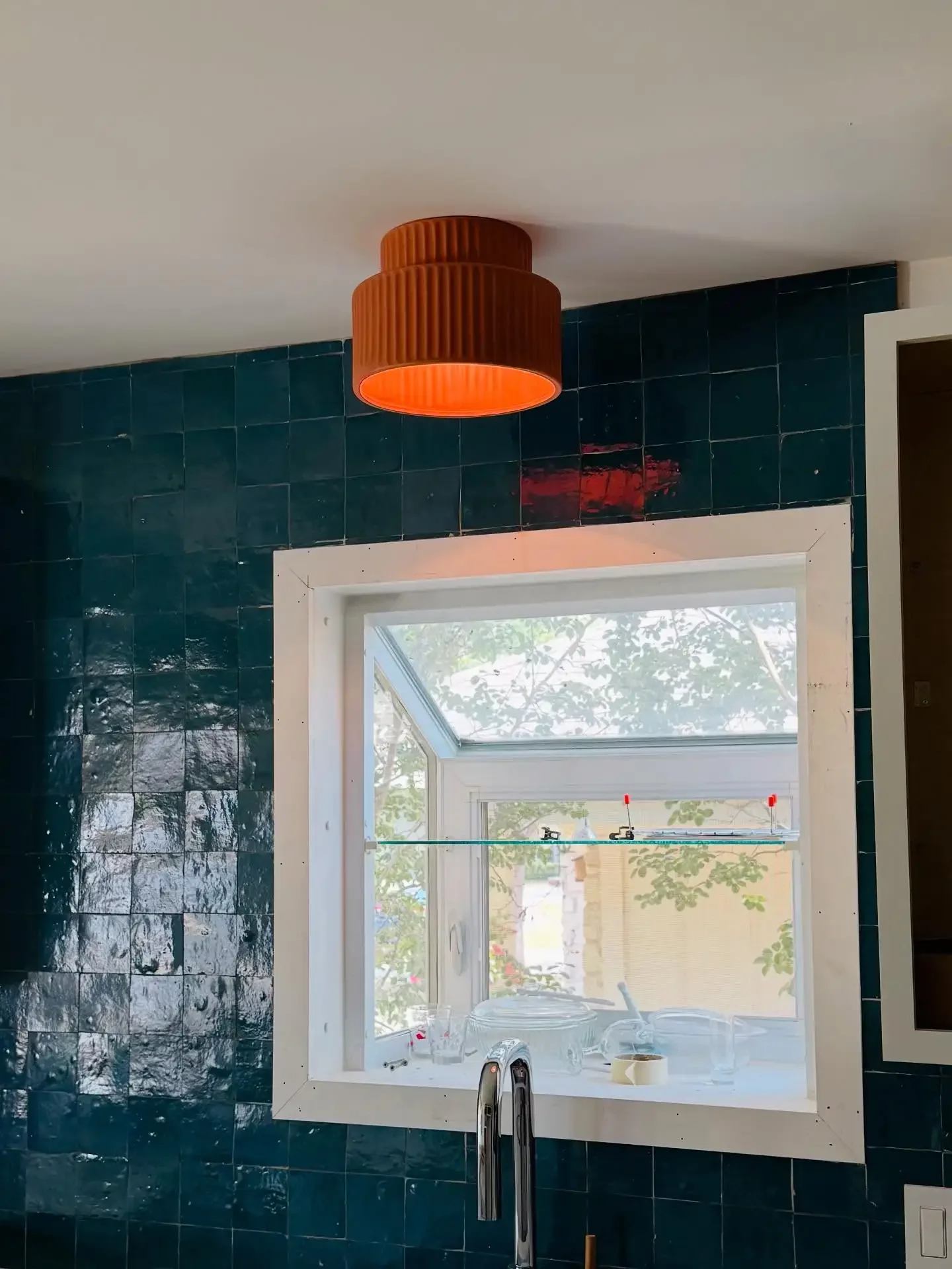 A kitchen with dark green tiled wall, a white-framed window showing outdoor greenery, and a modern copper-colored ceiling light fixture.