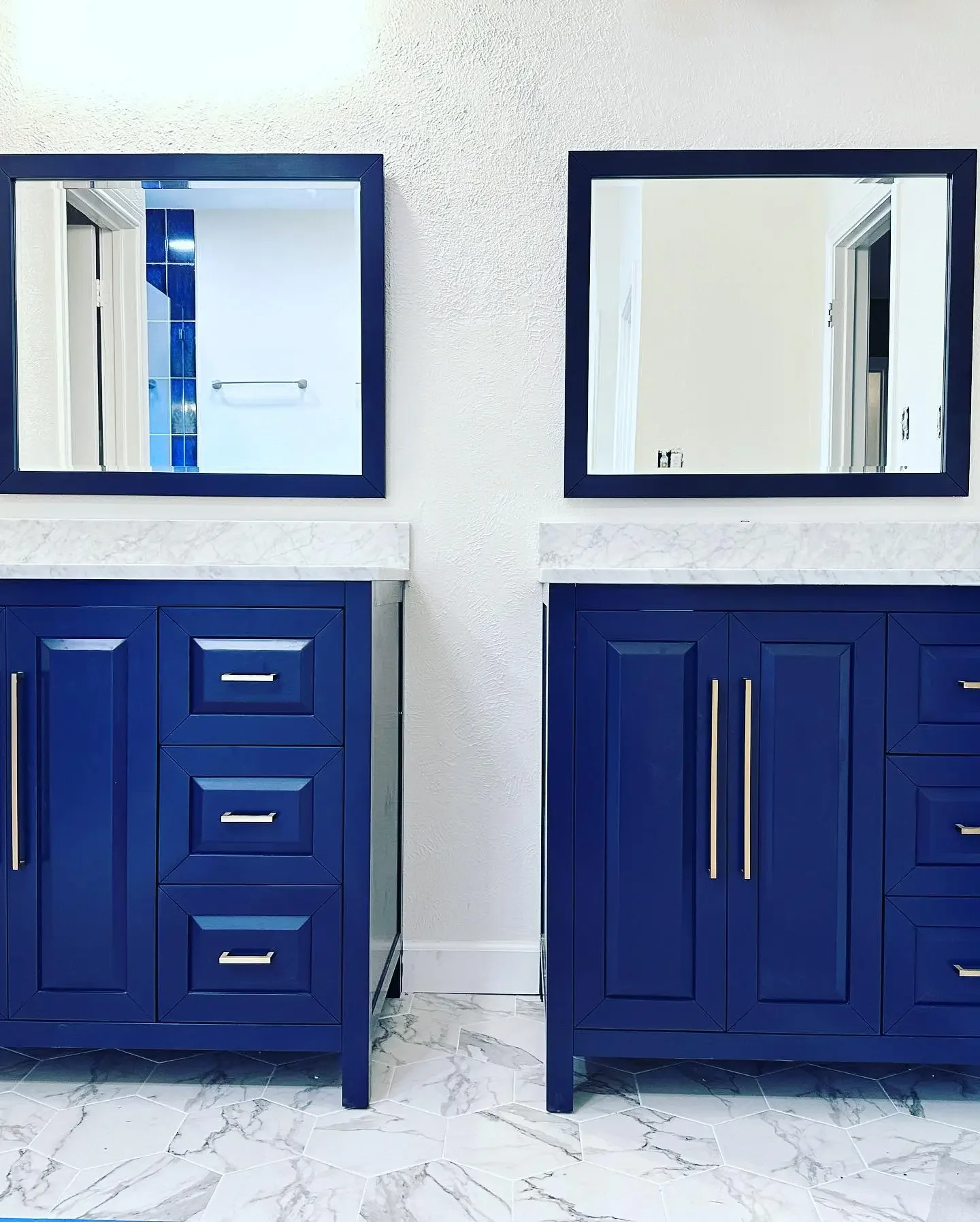 Two blue bathroom vanities with white marble countertops, each with a mirror above and gold handles, set against a white wall with hexagonal marble floor tiles.