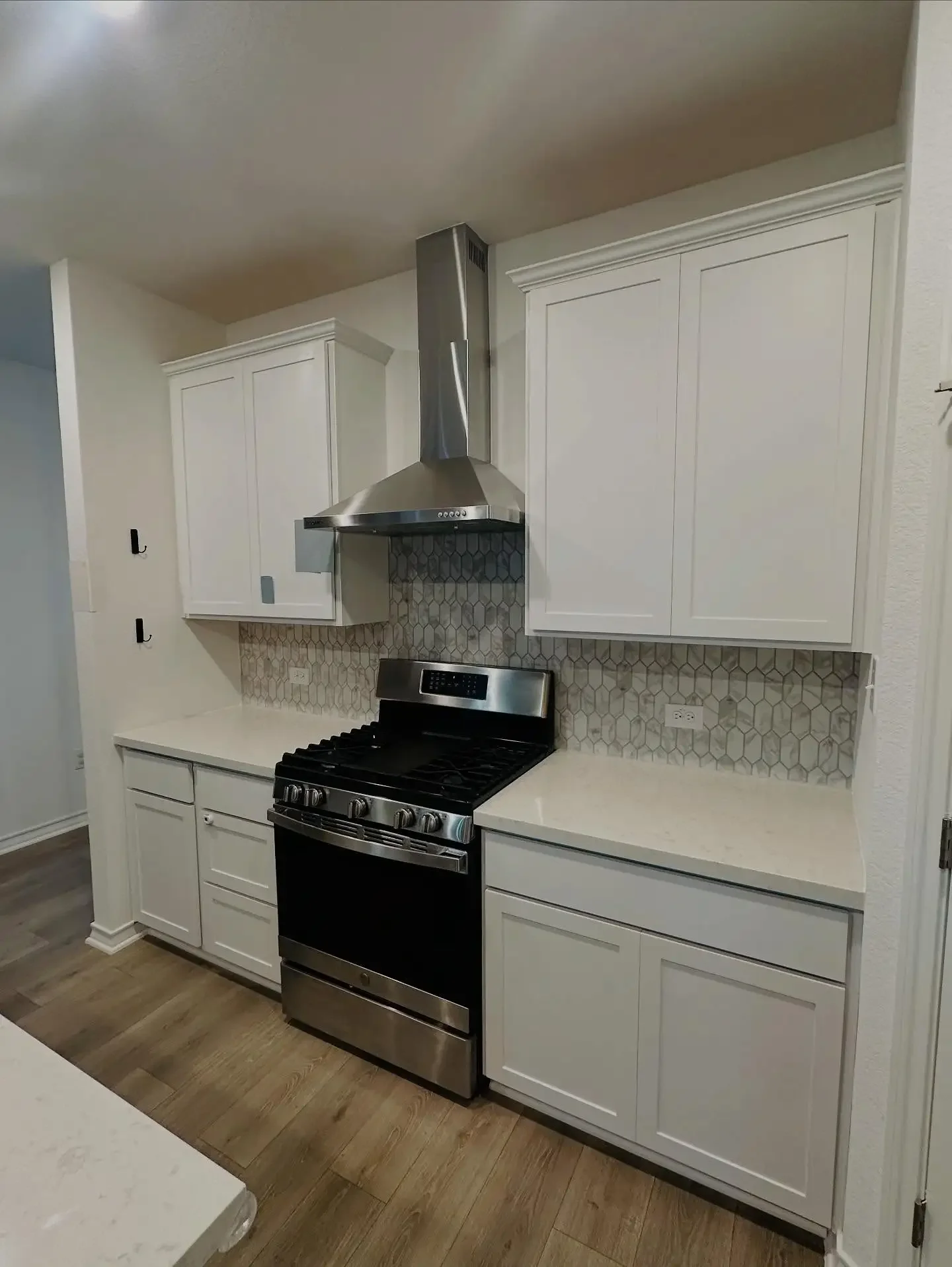 Kitchen with white cabinets, stainless steel oven, stove, and range hood, beige and gray hexagonal backsplash, and brown wood flooring.