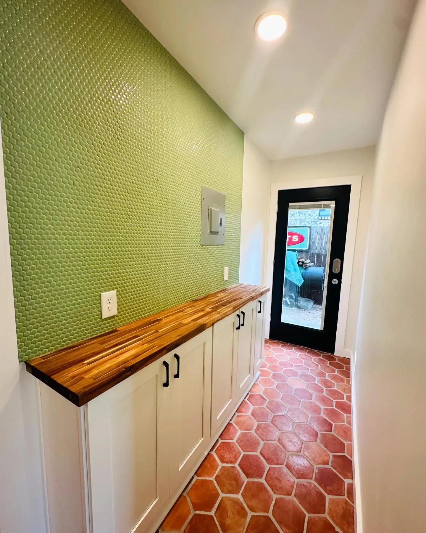 Kitchen or entryway with hexagonal terracotta tiles, white cabinets, green textured wall, wooden countertop, white walls, ceiling lights, glass door leading outside.