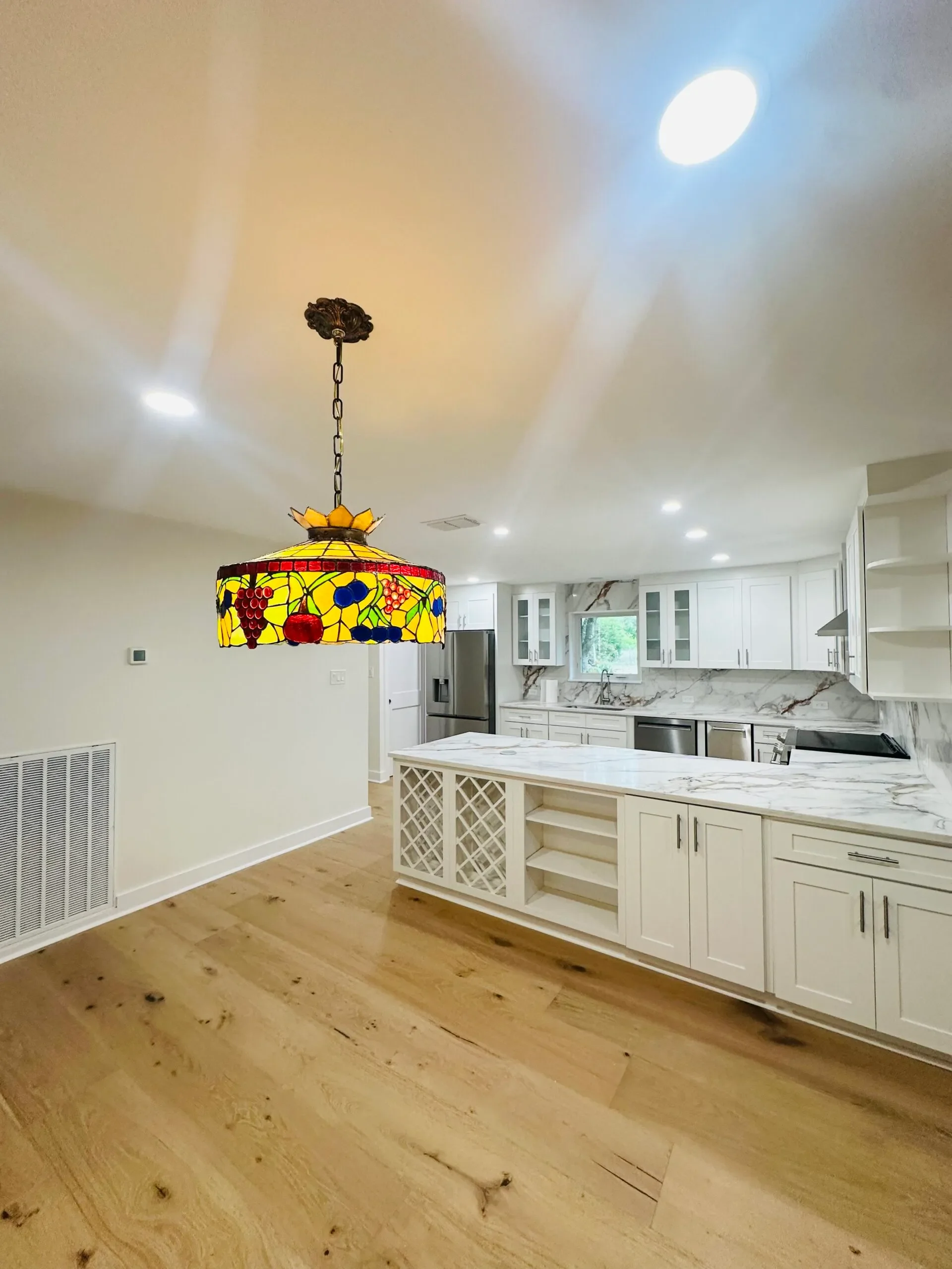 Bright kitchen with white cabinets, marble backsplash, and a central island. A colorful stained glass pendant light hangs from the ceiling, and natural light enters through a window.