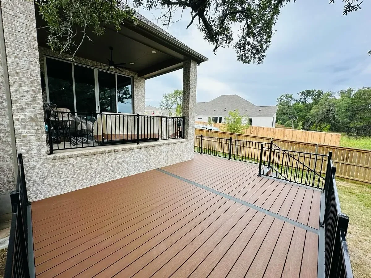 Large wooden deck with black metal railings attached to a house, showing a covered patio with ceiling fan and furniture, overlooking a backyard with a wooden fence and neighboring houses in the distance under a partly cloudy sky.
