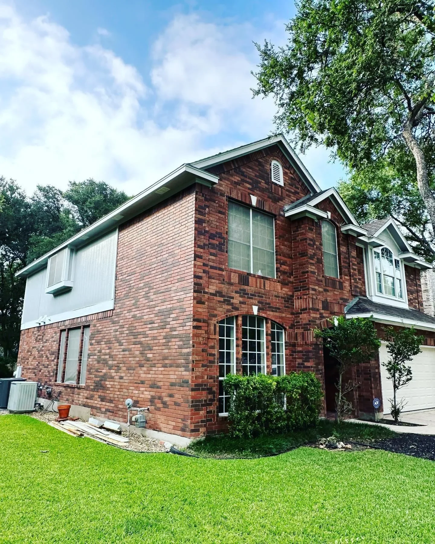 A two-story brick house with a white garage door, large windows, and a landscaped front yard with green grass and bushes, under a partly cloudy sky.