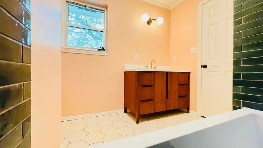 Bathroom with peach walls, a window, a wooden vanity with a sink, a wall-mounted light fixture, white hexagon tile flooring, a green tiled wall, and a white door.
