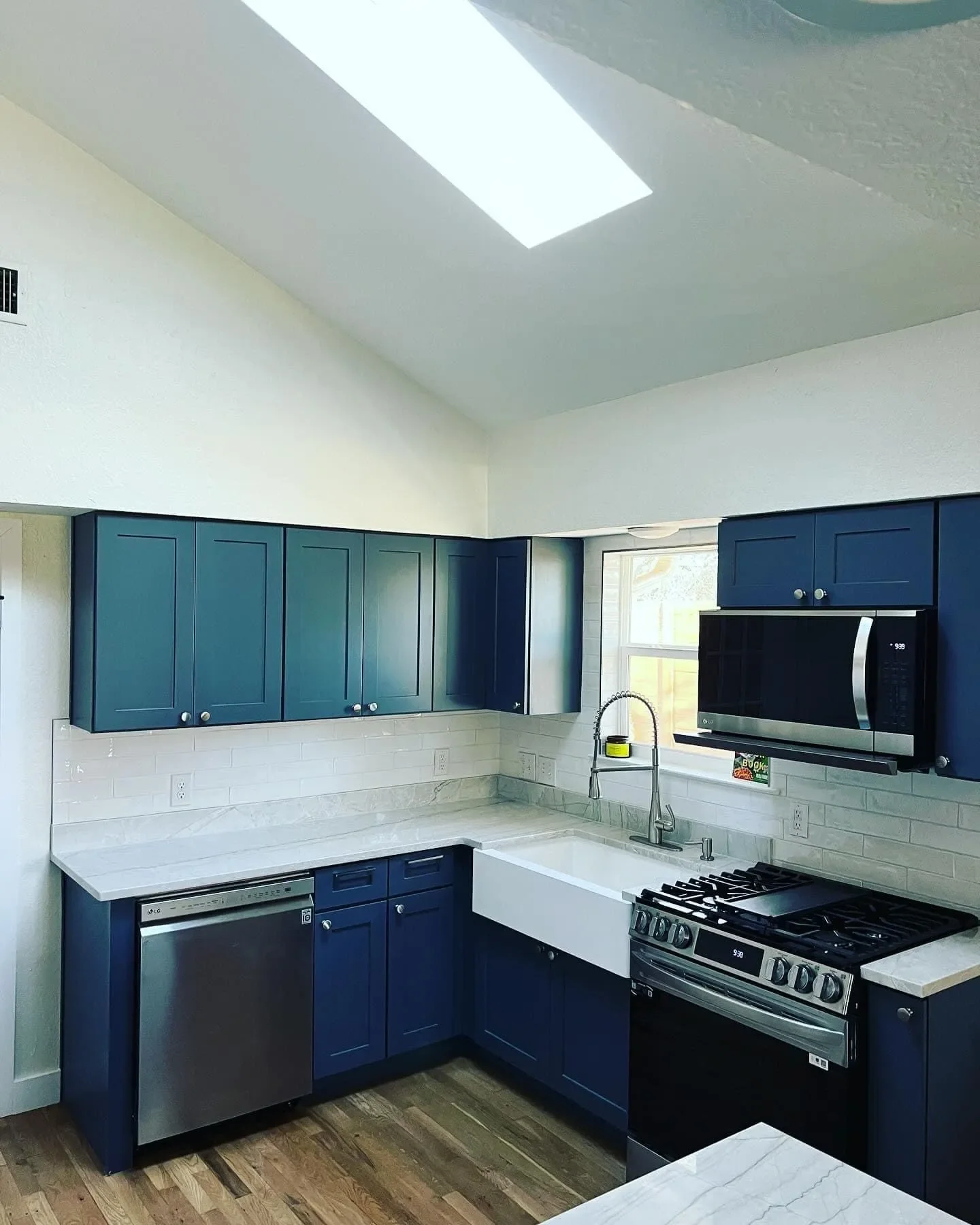 Kitchen with blue upper and lower cabinets, white marble countertops, a farmhouse sink, stainless steel dishwasher and gas stove, microwave, wooden floor, and a small window above the sink.