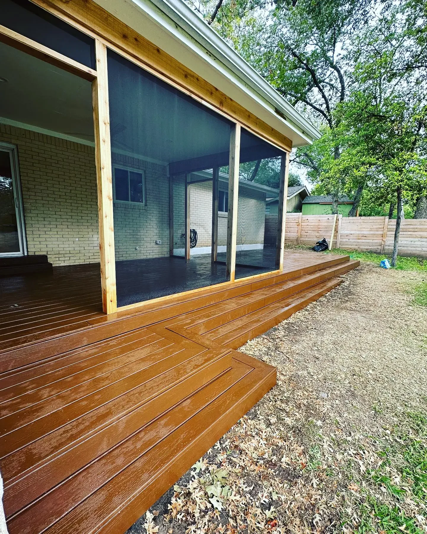Newly built wooden deck with a screened-in porch attached to a brick house, with trees and a wooden fence in the background.