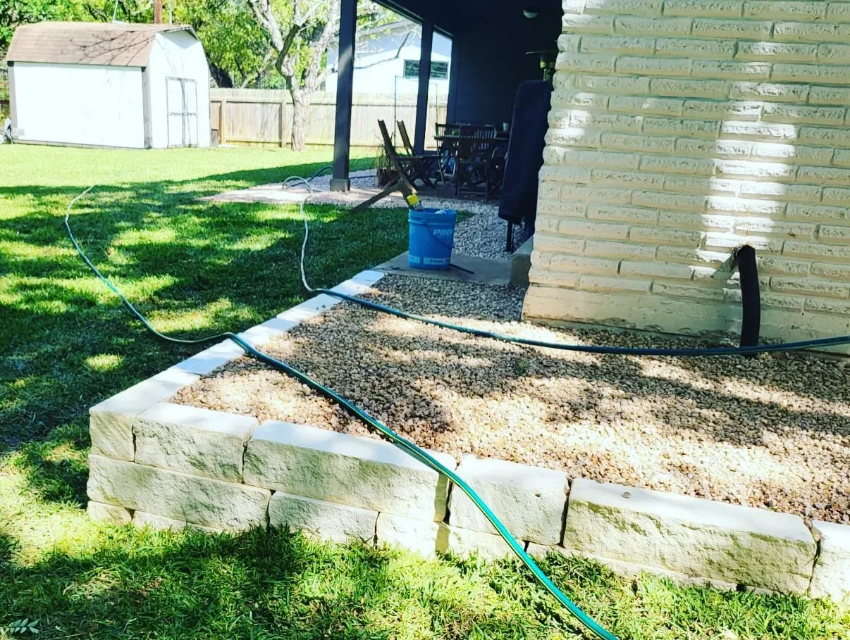 Backyard with a stone-bordered garden bed, a black hose, and a blue bucket. There's a shed and a fenced yard with trees in the background.